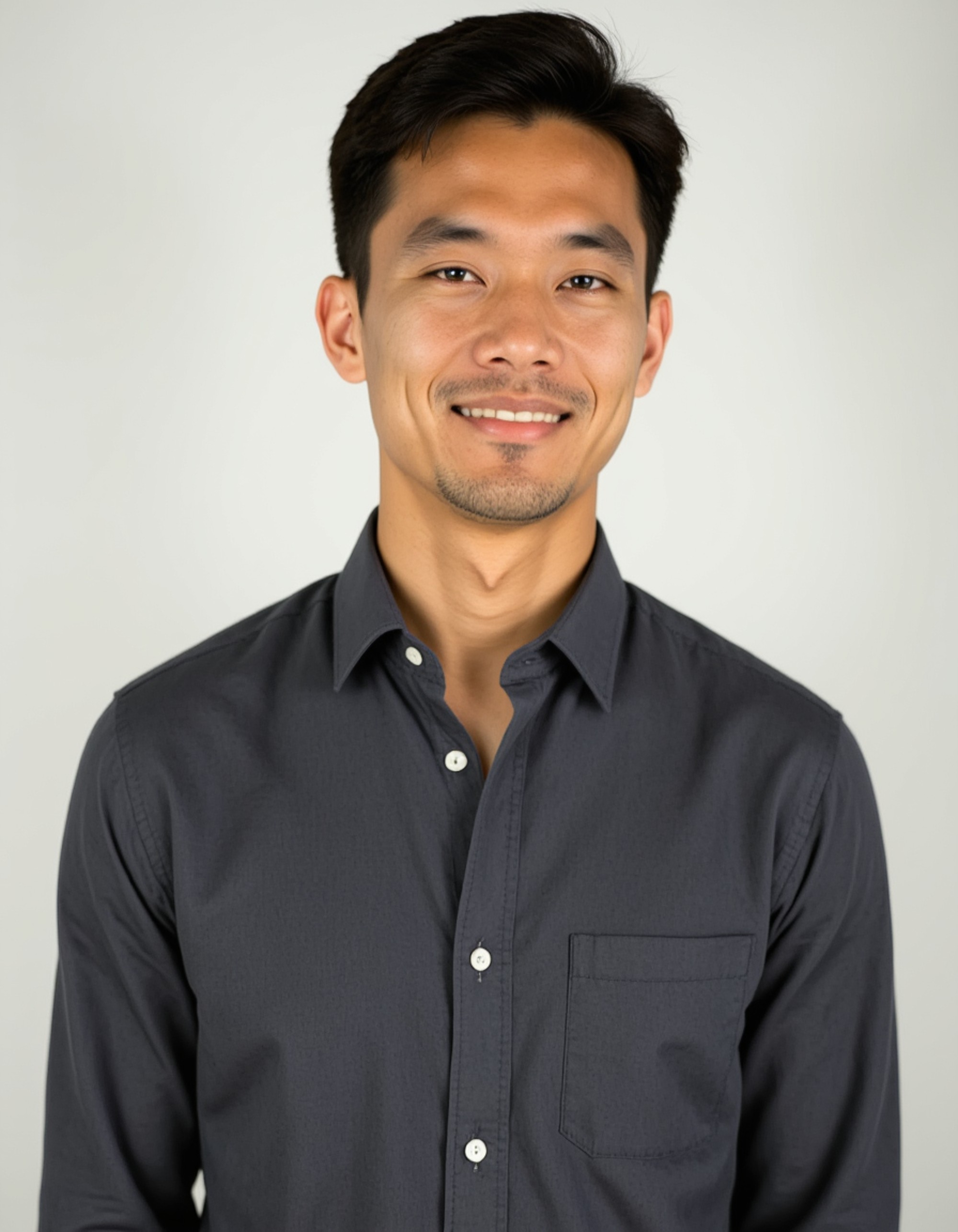 A professional actor headshot of a confident male model, taken in a well-lit studio with a clean white backdrop. The model is wearing a charcoal gray button-down shirt with the top button undone and sleeves rolled up slightly. His dark hair is styled with a modern side part and he has a well-groomed beard. The lighting is soft and even, highlighting his strong jawline and creating catchlights in his eyes with a relaxed, genuine smile perfect for leading man roles.
